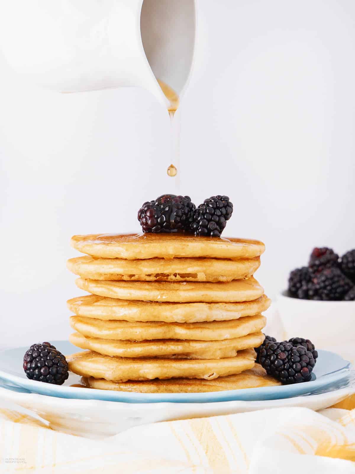 A stack of pancakes topped with fresh blackberries on a blue plate. Syrup is being poured over them from a white pitcher. A bowl of blackberries is in the background on a light-colored tablecloth.