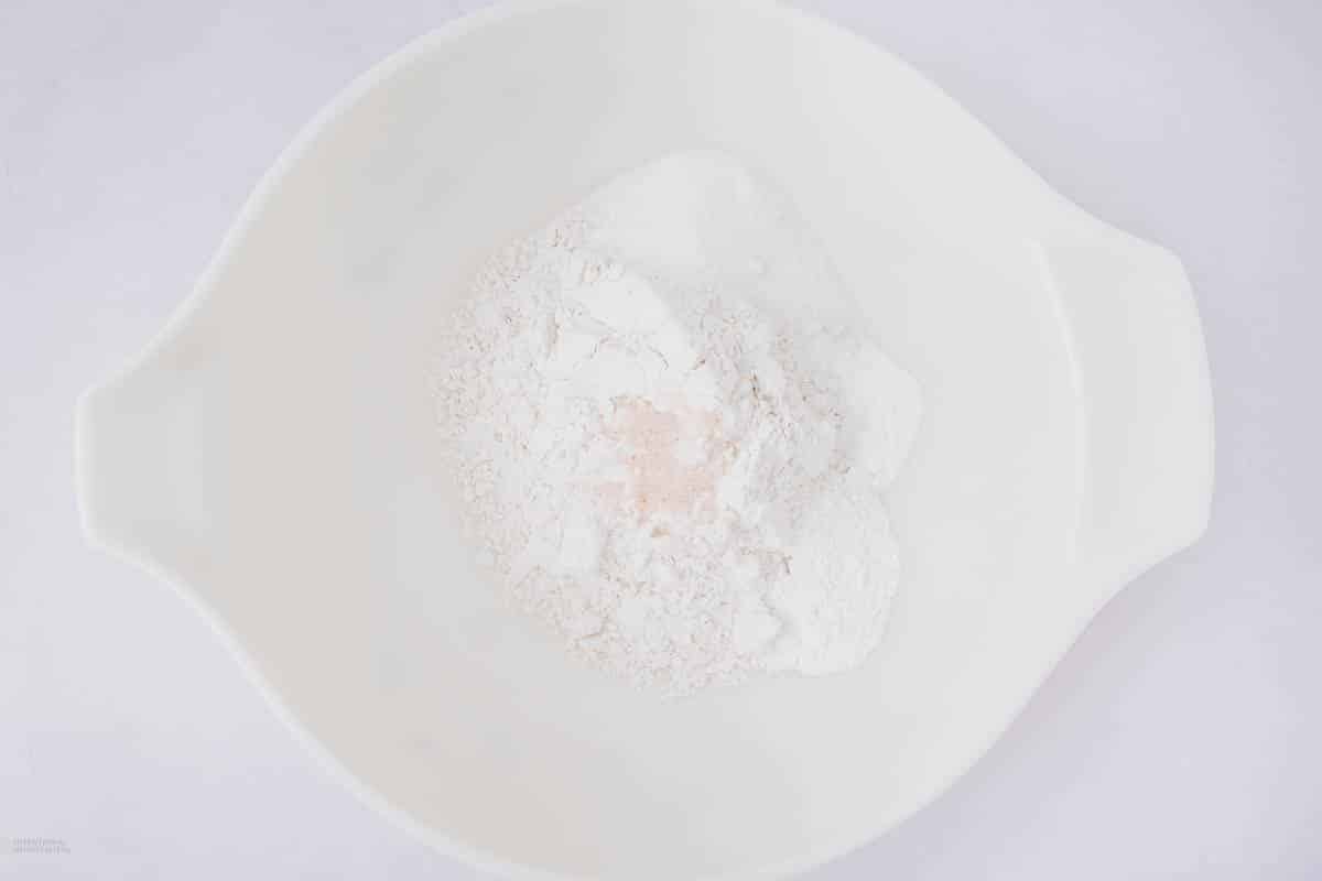 A white mixing bowl containing various dry baking ingredients, including flour, sugar, and salt, on a light-colored surface.