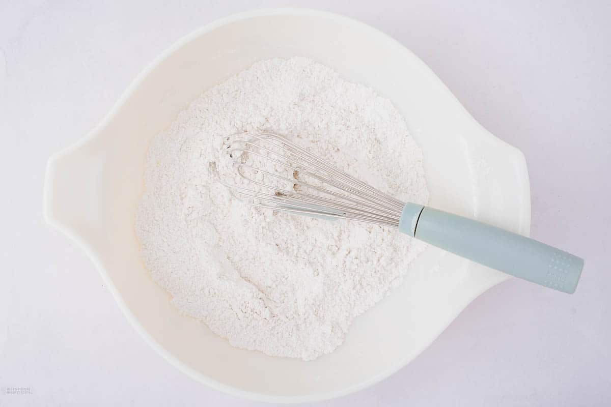 A white mixing bowl containing flour and a whisk with a blue handle resting on top, set against a light background.