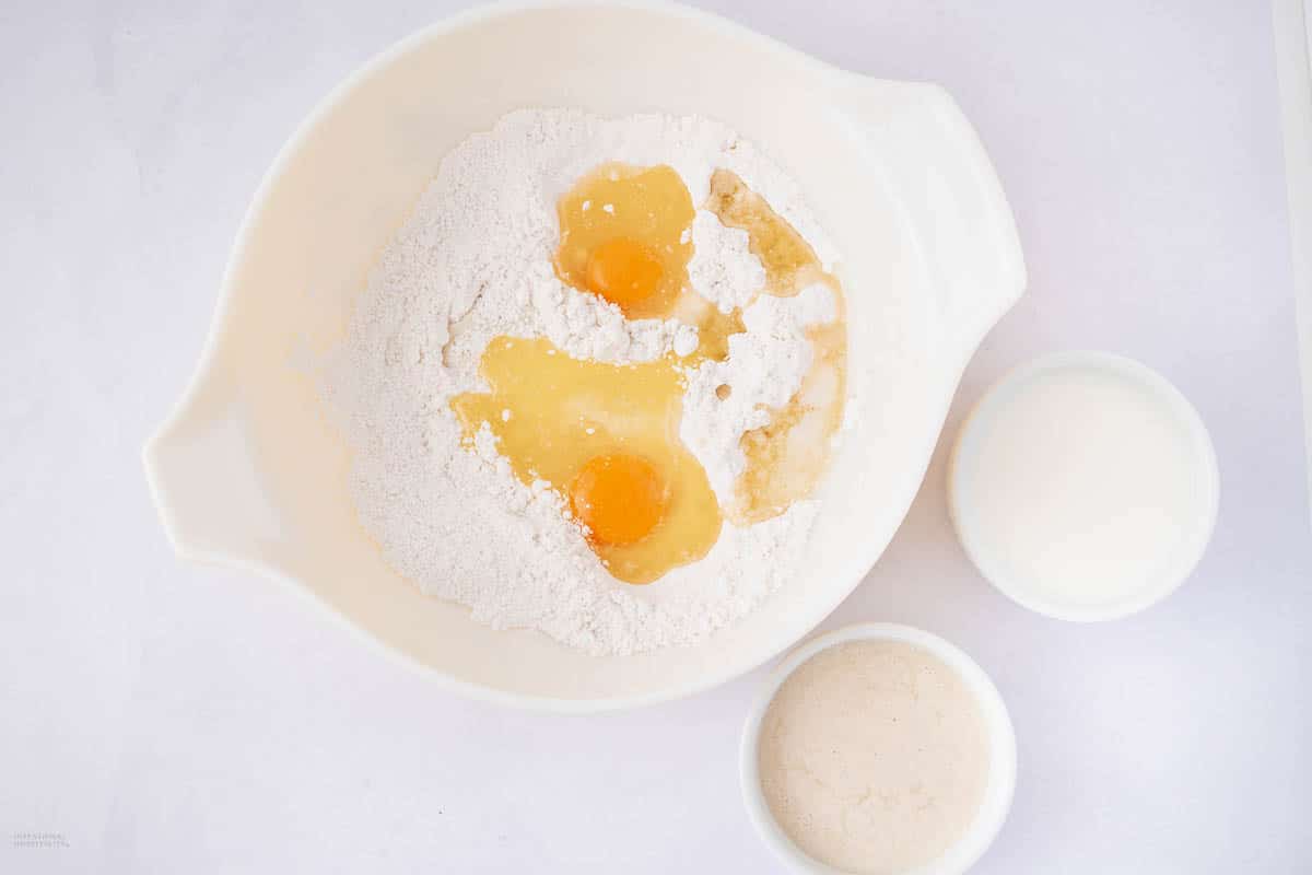 A mixing bowl filled with flour and two cracked eggs on a white surface. Next to the bowl are two smaller bowls, one with a liquid ingredient and the other with a creamy mixture, all prepared for baking.