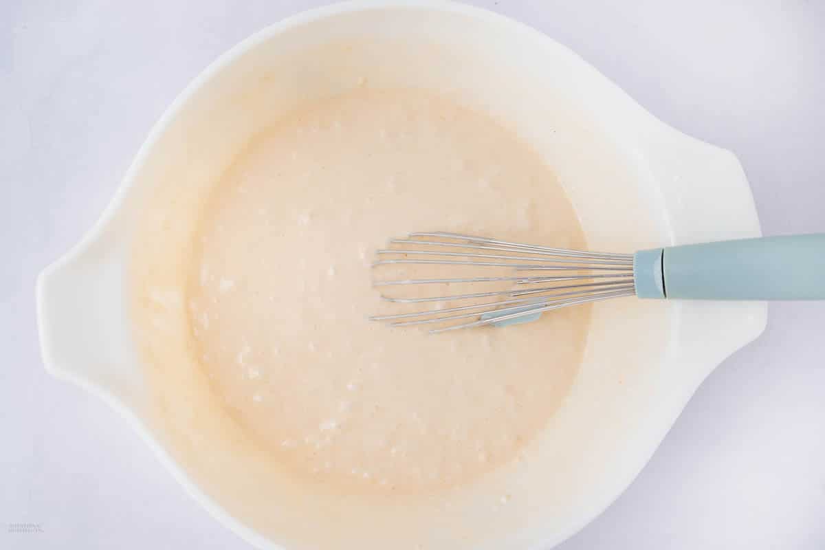 A light beige batter in a white mixing bowl with a blue-handled whisk resting on the side, set against a plain white background.