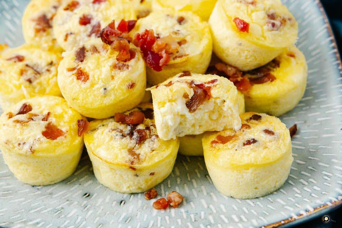 A pile of fluffy egg bites topped with bits of bacon on a textured plate. One bite is cut in half, revealing a creamy interior. The bites are golden and appear moist and appetizing.