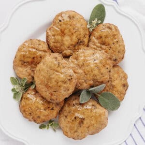 A white plate with a pile of browned sausage patties garnished with fresh sage leaves. The patties appear juicy and are glistening, suggesting they have just been cooked. A striped cloth is partially visible underneath the plate.