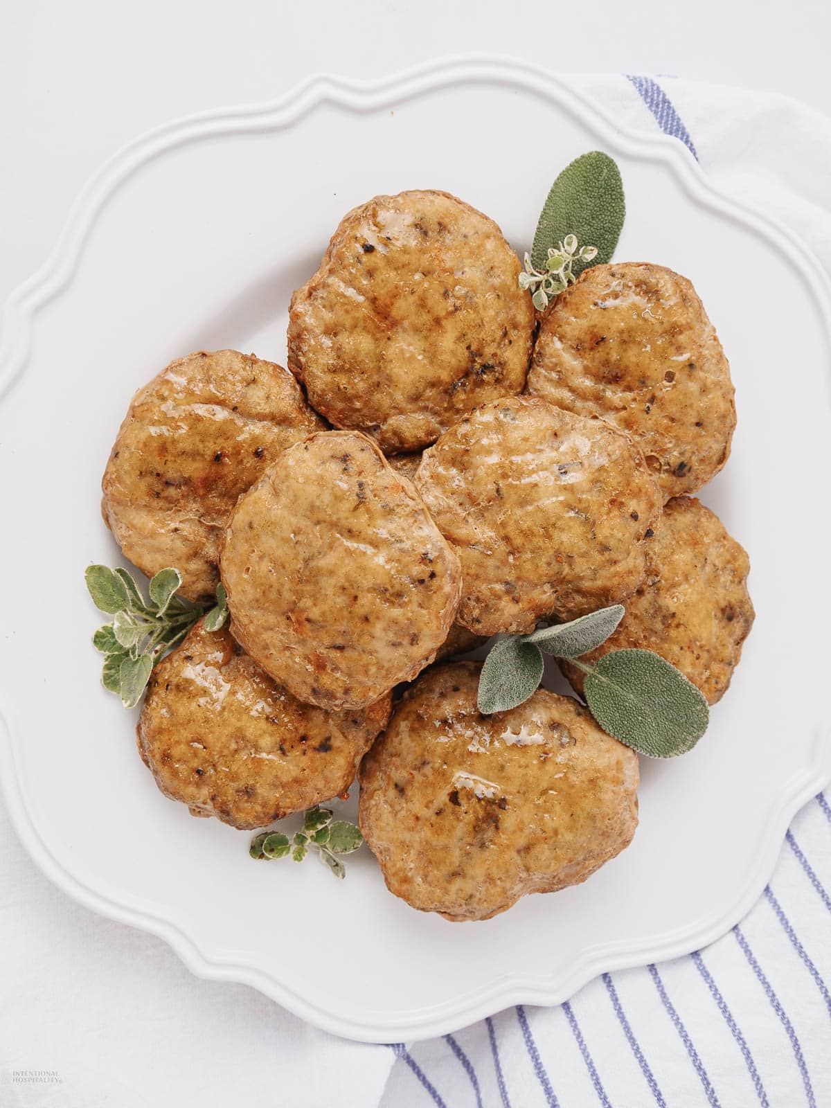 A white plate with eight browned sausage patties garnished with fresh sage and oregano leaves. The plate is placed on a white and blue striped cloth.