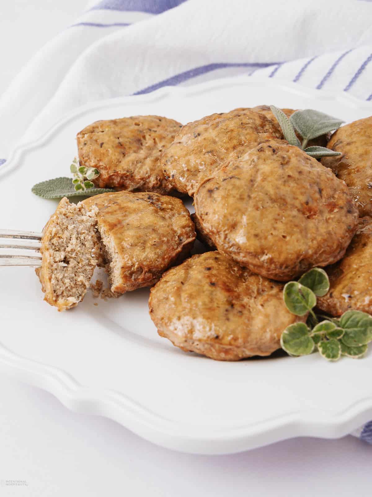 A white plate with several browned sausage patties garnished with fresh herbs. One patty is cut in half with a fork resting nearby, revealing a juicy interior. A striped cloth is partially visible in the background.