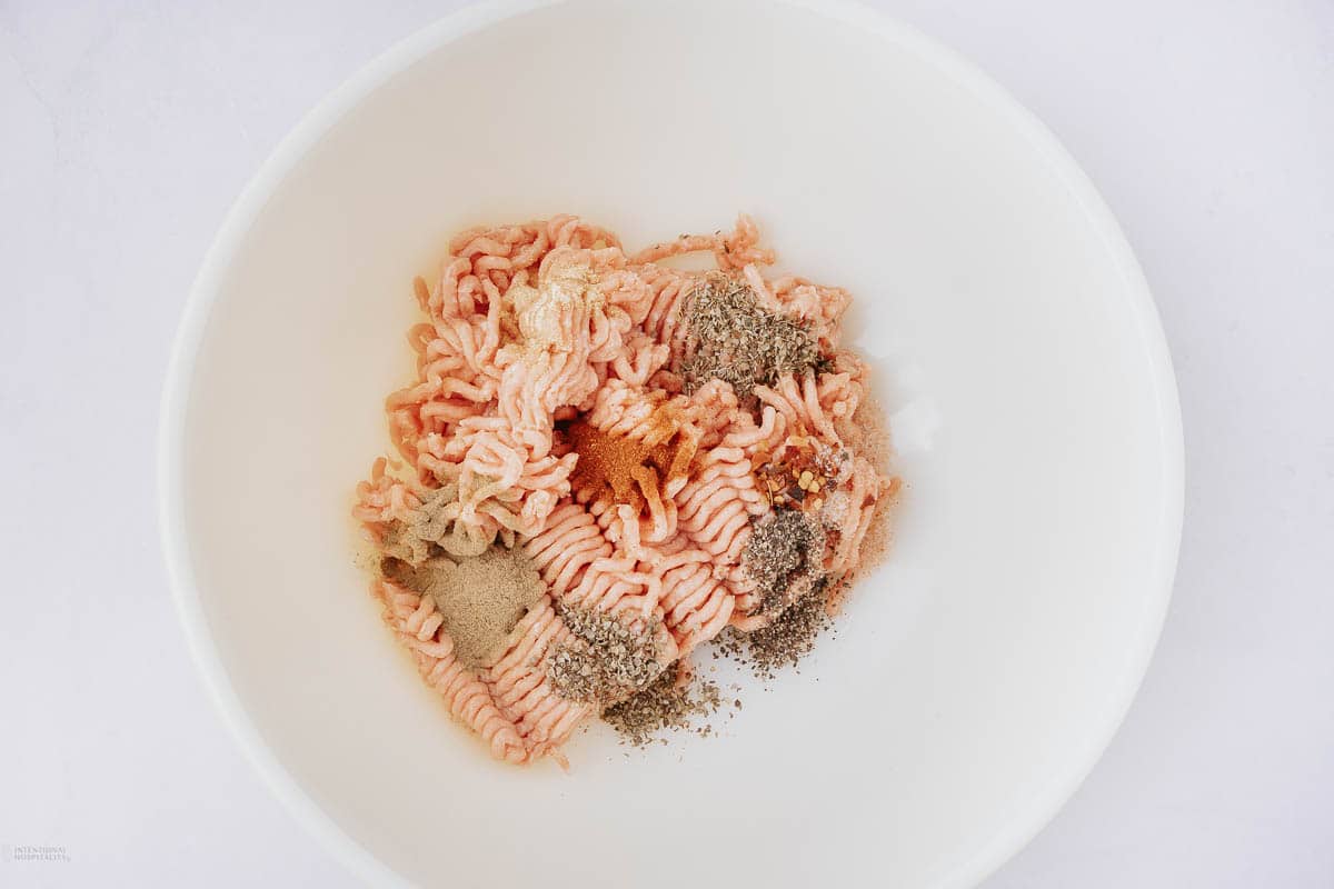 A white bowl filled with raw ground meat mixed with various spices and seasonings, including black pepper and a reddish-brown spice. The ingredients are ready to be mixed together for a recipe.