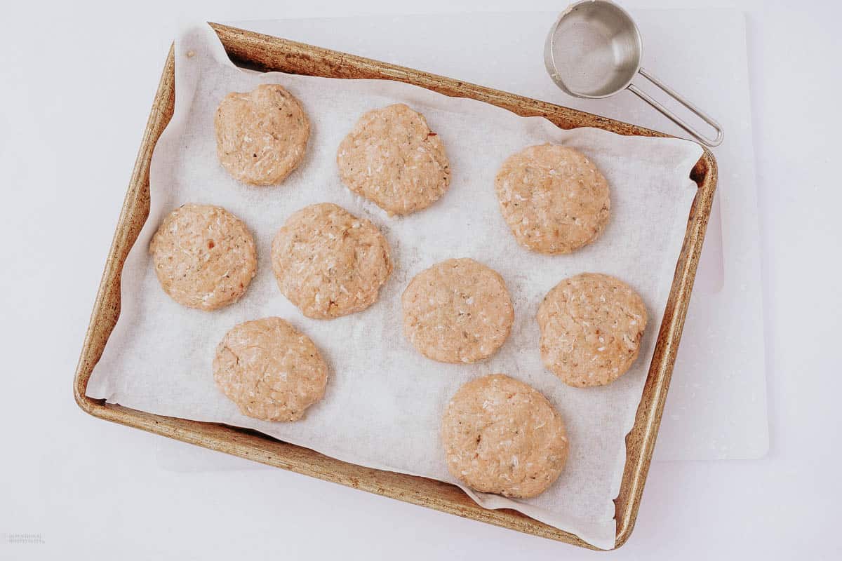 A baking tray lined with parchment paper holds eight raw, round biscuit dough portions. A metal measuring cup is placed to the right of the tray on a white surface.