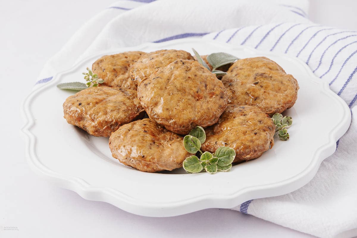 Seven cooked sausage patties are arranged on a white plate, garnished with fresh herbs. A white and blue striped cloth is underneath the plate.