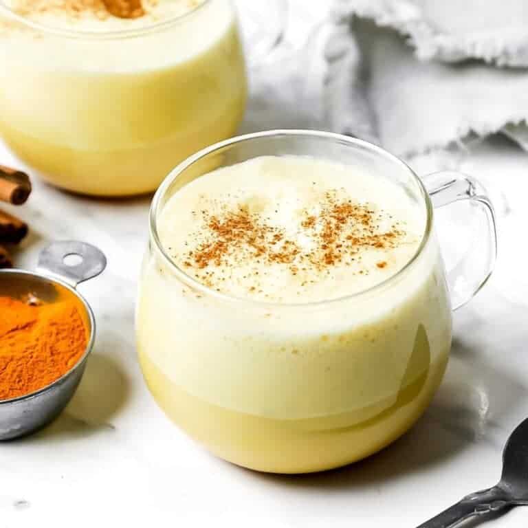 A glass mug filled with frothy golden milk, sprinkled with cinnamon, sits on a white marble surface. A small dish with golden powder is nearby, and a beige cloth is in the background. Another similar mug is partially visible.