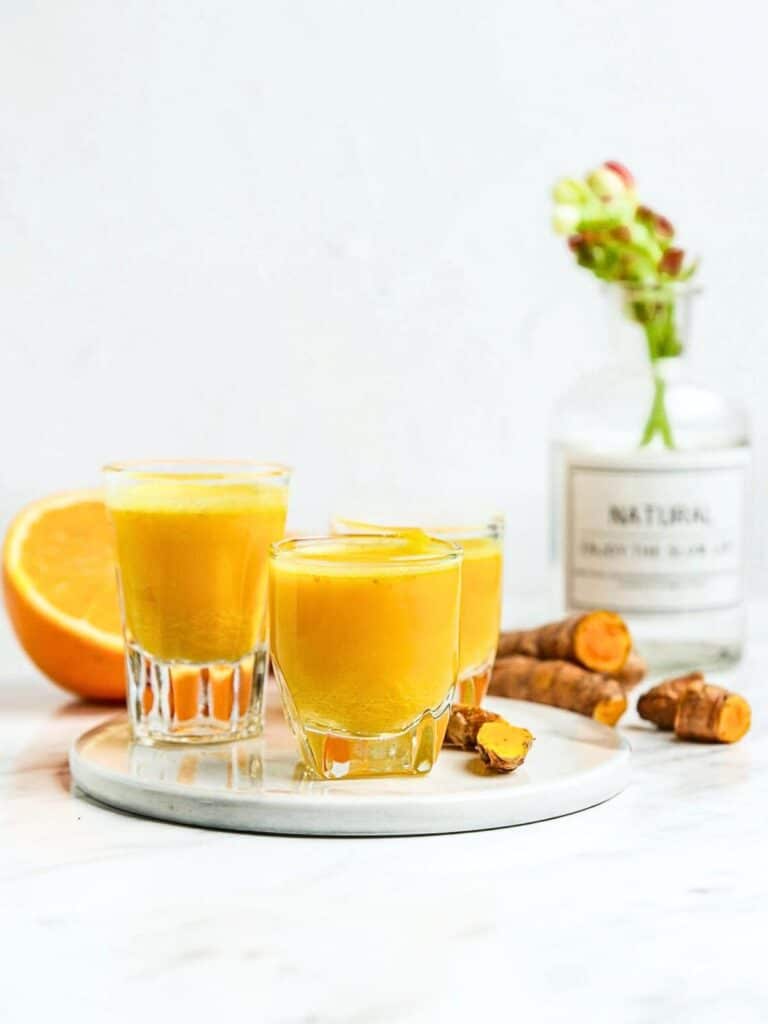 Three small glasses filled with bright yellow turmeric shots sit on a round white plate. Fresh turmeric roots are scattered around, with half an orange beside them. A plant and a labeled bottle are in the blurred background.