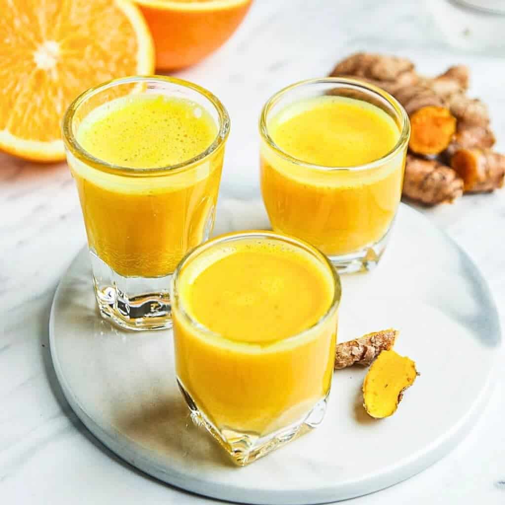 Three small glasses filled with bright orange turmeric shots are placed on a round white plate. A sliced orange and pieces of fresh turmeric root are visible in the background. The setting is on a light marble surface.