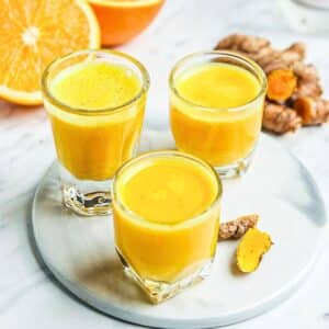 Three small glasses filled with bright orange turmeric shots are placed on a round white plate. A sliced orange and pieces of fresh turmeric root are visible in the background. The setting is on a light marble surface.