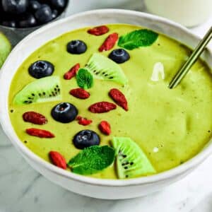 A vibrant green smoothie bowl topped with fresh kiwi slices, blueberries, goji berries, and mint leaves. A metal spoon is positioned on the right, and a bowl of blueberries is visible in the background on a marble surface.