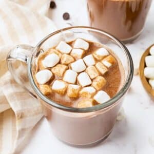 A clear glass mug filled with hot chocolate topped with mini marshmallows sits on a marble surface. A beige striped cloth is partially visible beside the mug, with another cup and a small bowl of marshmallows in the background.