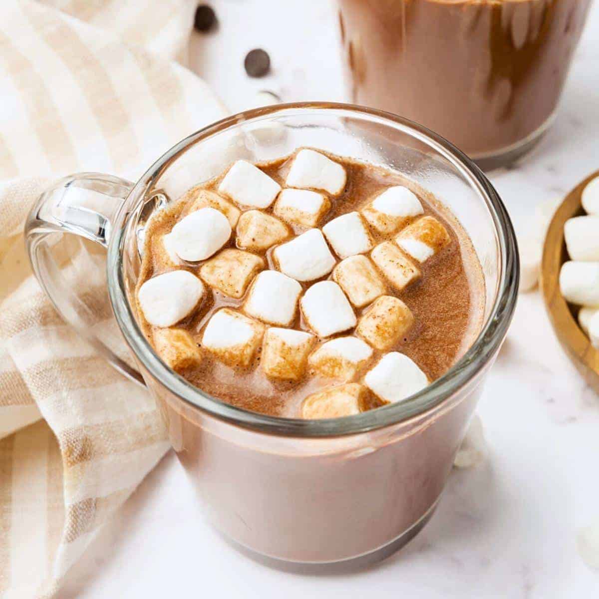 Hot chocolate with marshmallows on top in a glass mug on a white surface, a dish towel next to it