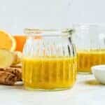 A clear glass jar filled with a vibrant yellow smoothie sits on a light surface. Behind it, there are slices of fresh orange and ginger with another jar partially visible, suggesting a healthy drink arrangement.
