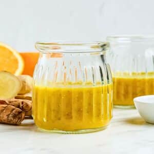 A clear glass jar filled with a vibrant yellow smoothie sits on a light surface. Behind it, there are slices of fresh orange and ginger with another jar partially visible, suggesting a healthy drink arrangement.