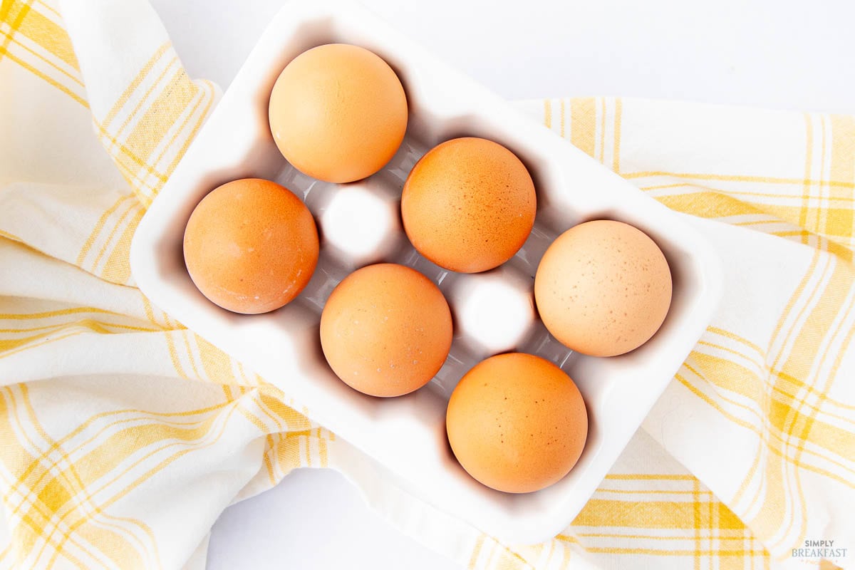 A carton of six brown eggs rests on a white surface, accompanied by a white and yellow checkered cloth.