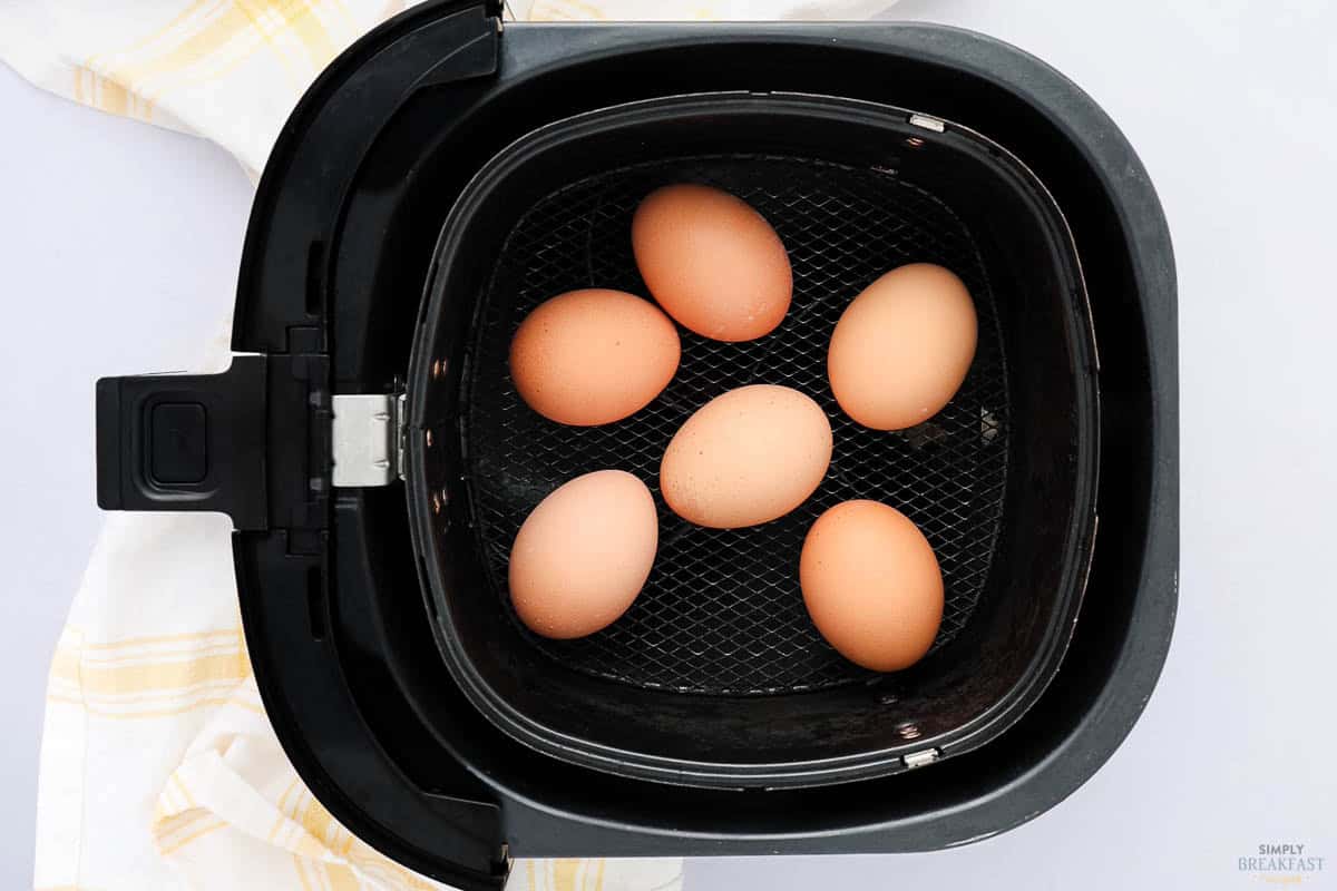 Five brown eggs inside an air fryer basket, with a yellow and white checkered cloth in the background.