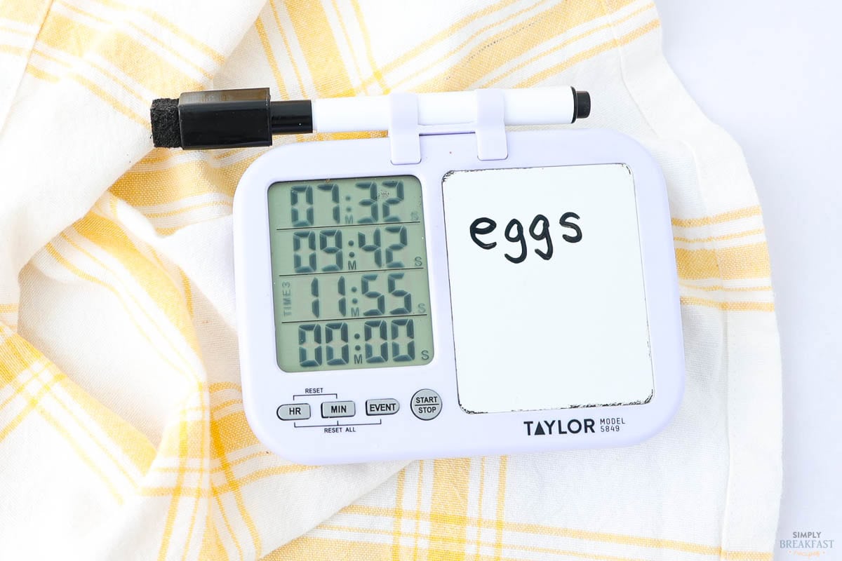 A digital kitchen timer displays times 07:32, 09:42, 11:55, and 01:00, with an attached small whiteboard. The word "eggs" is written on the whiteboard. A black marker is clipped on top. A yellow plaid cloth is partially visible.