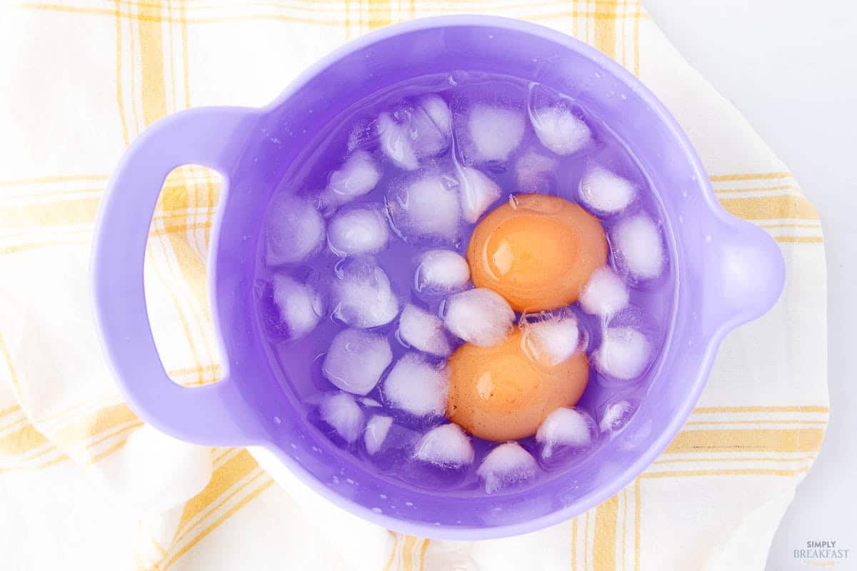 Two brown eggs in a purple bowl filled with ice water and ice cubes. The bowl is placed on a yellow and white checkered cloth.