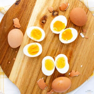 Top view of a round wooden board with four boiled eggs, two peeled and halved, displaying bright yolks—some soft, runny, and others firm. Eggshells scattered around, with two uncracked eggs nearby. A yellow and white cloth partially visible.