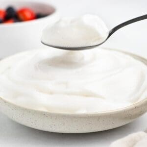 A spoonful of creamy yogurt being lifted from a bowl filled with yogurt. In the blurred background, there's a bowl containing berries.