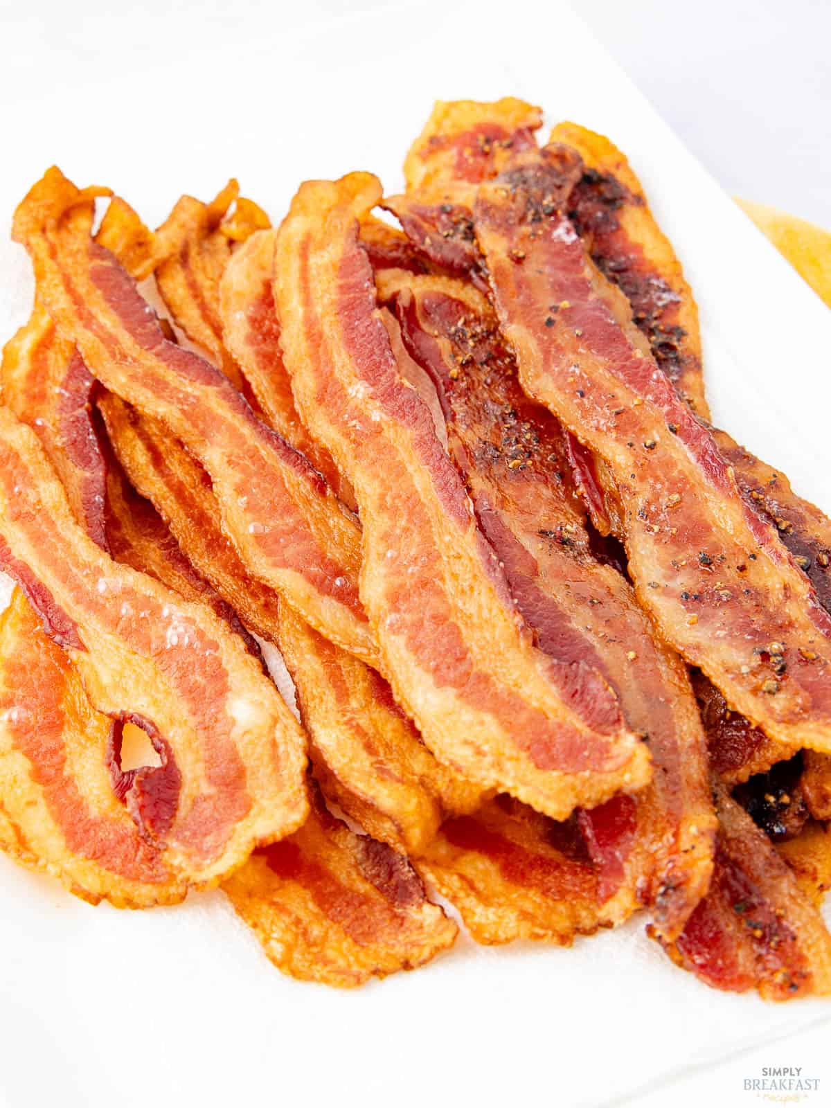 Crispy strips of cooked bacon stacked on a white paper towel, showcasing a golden-brown texture with edges slightly curled. Perfect results are easy when you know how to make bacon in the oven—each piece freshly cooked, some showing darker, caramelized areas.