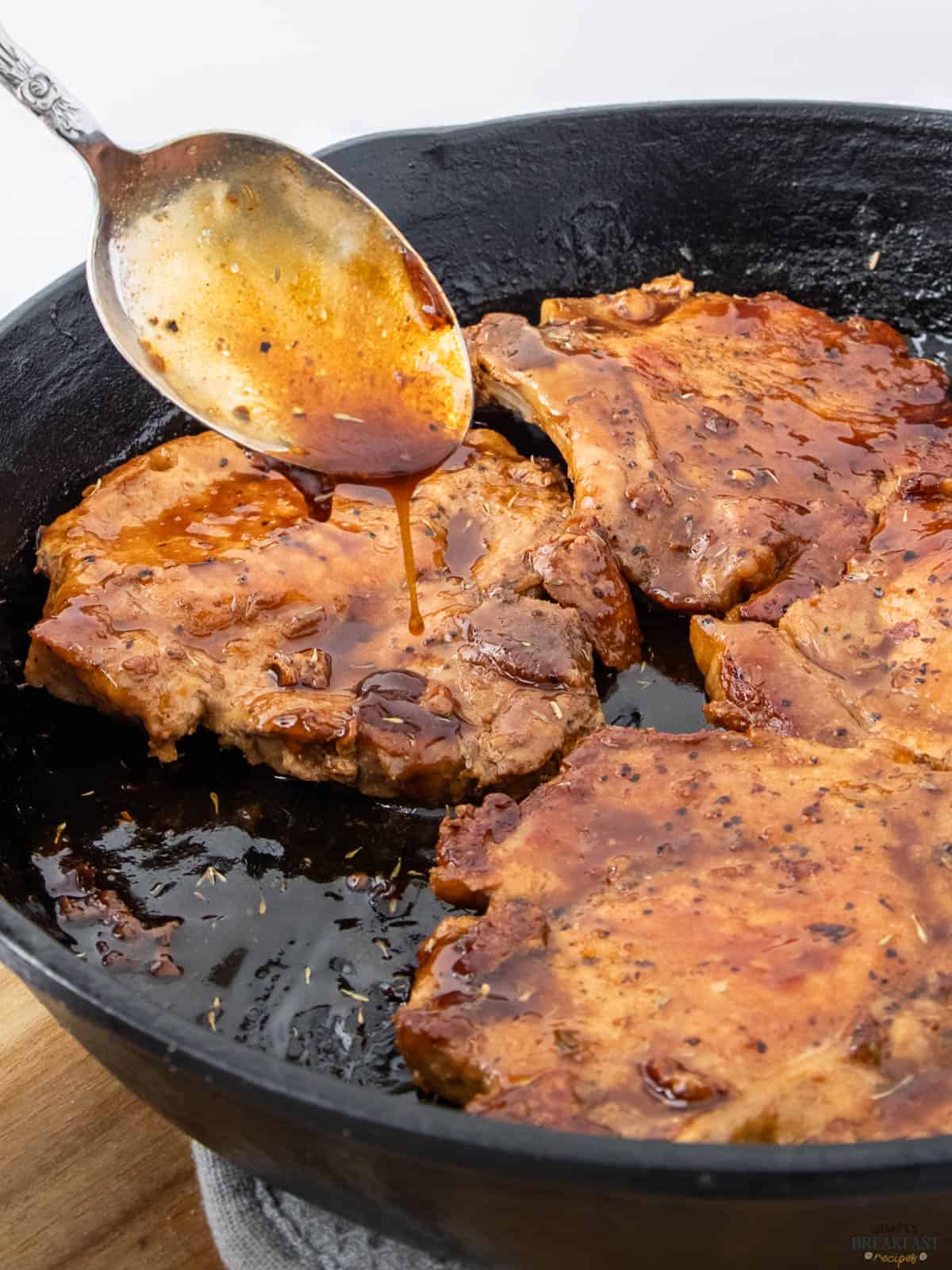 A spoon drizzles sauce over browned pork chops in a black skillet. The meat is evenly cooked, and the sauce is glossy, adding a savory appearance. The seasoned cast iron skillet enhances the dish's rustic presentation.