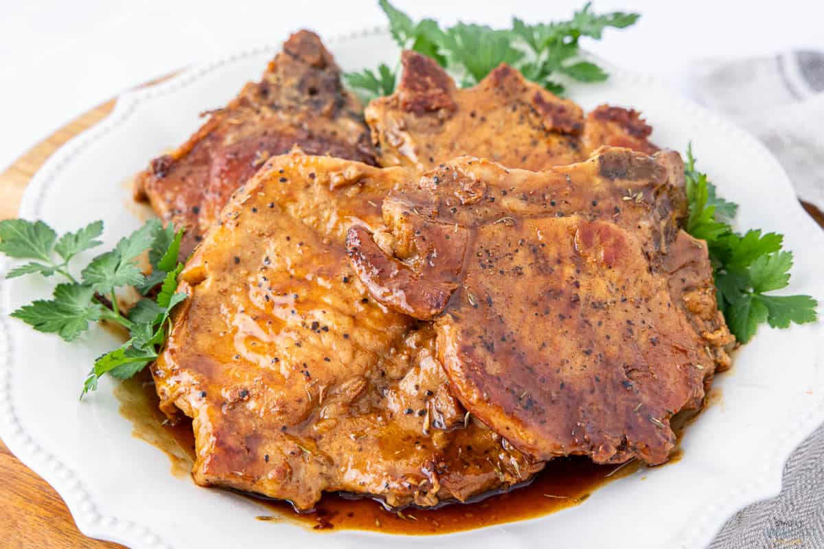 Cooked pork chops glazed in a rich sauce sit on a white plate, garnished with fresh parsley on the sides. The sauce glistens, highlighting the savory seasoning on the meat.