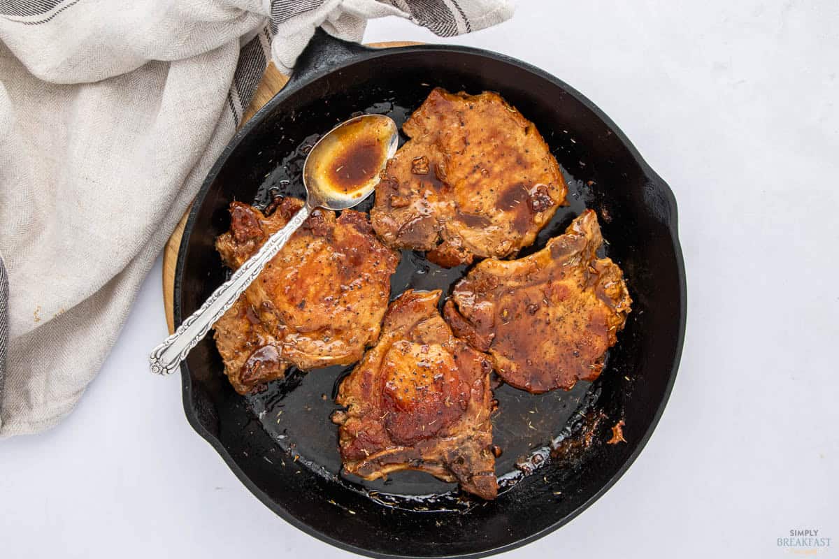 A black skillet contains four cooked pork chops, glazed with a dark sauce. A silver spoon rests in the sauce at the side. A striped cloth napkin partially covers the skillet handle on a light surface.