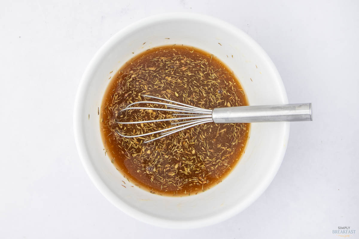 A white bowl is filled with a brown sauce containing herbs. A metal whisk rests inside the bowl, suggesting mixing. The background is a smooth, white surface.