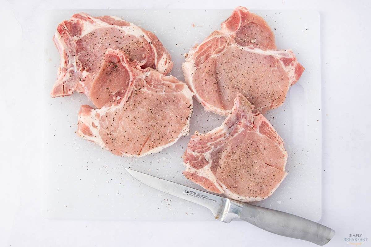 Four raw pork chops seasoned with pepper are on a white cutting board. A knife is placed beside the pork chops.