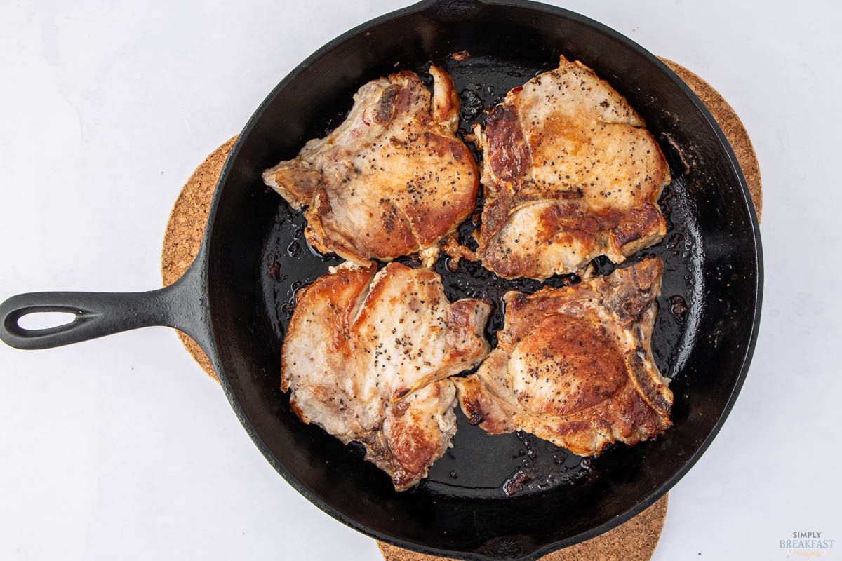 Four cooked pork chops seasoned with pepper are in a black cast iron skillet. The skillet rests on a round cork trivet against a light background.