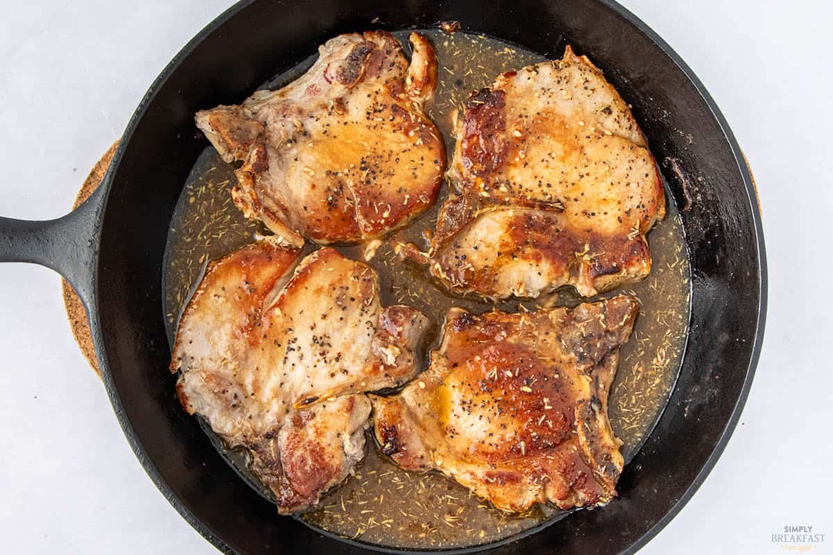 Four browned pork chops seasoned with pepper are cooking in a cast iron skillet. The pan contains a light sauce, and the meat is garnished with herbs. The background is a light surface.