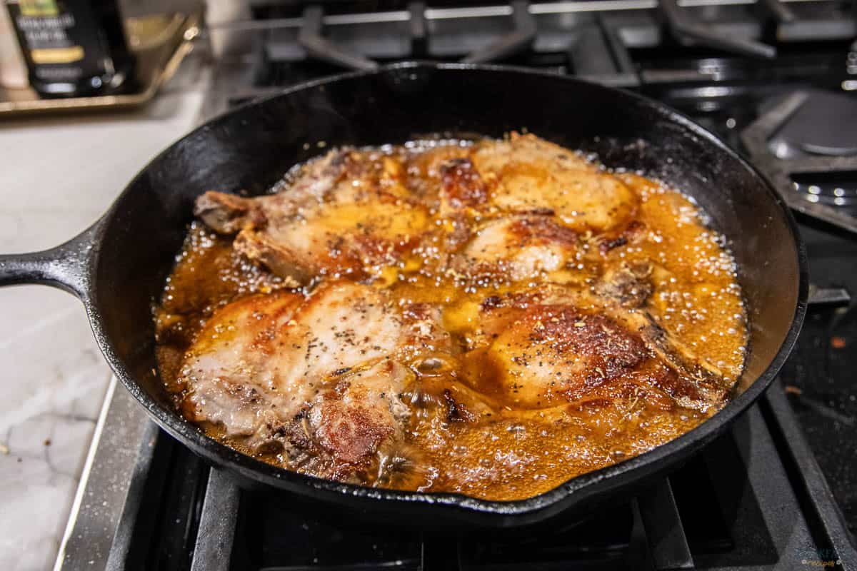 A cast iron skillet on a stove contains golden-brown pork chops sizzling in bubbling hot oil, seasoned with black pepper. The rich sauce surrounds the meat, indicating a savory and flavorful preparation.