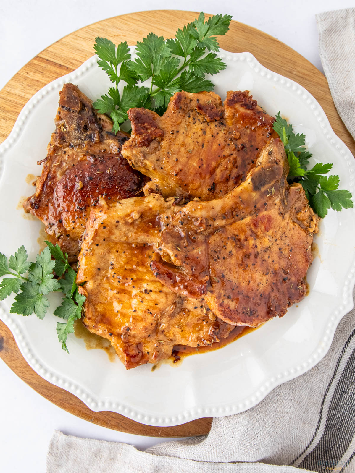 Plate of saucy, seasoned pork chops garnished with fresh parsley, set on a wooden board with a striped cloth napkin.