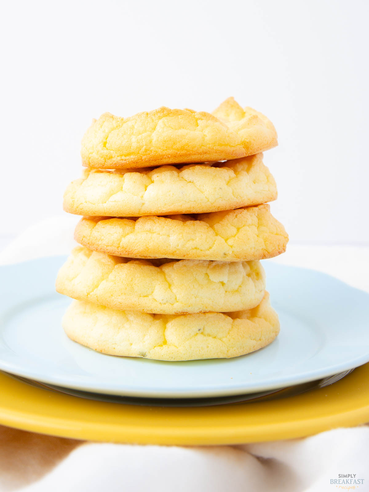 A stack of five light, fluffy cookies sits on a pale blue plate, which is placed on a yellow plate. The cookies have a slightly cracked surface and a golden-brown edge. The background is soft and neutral.