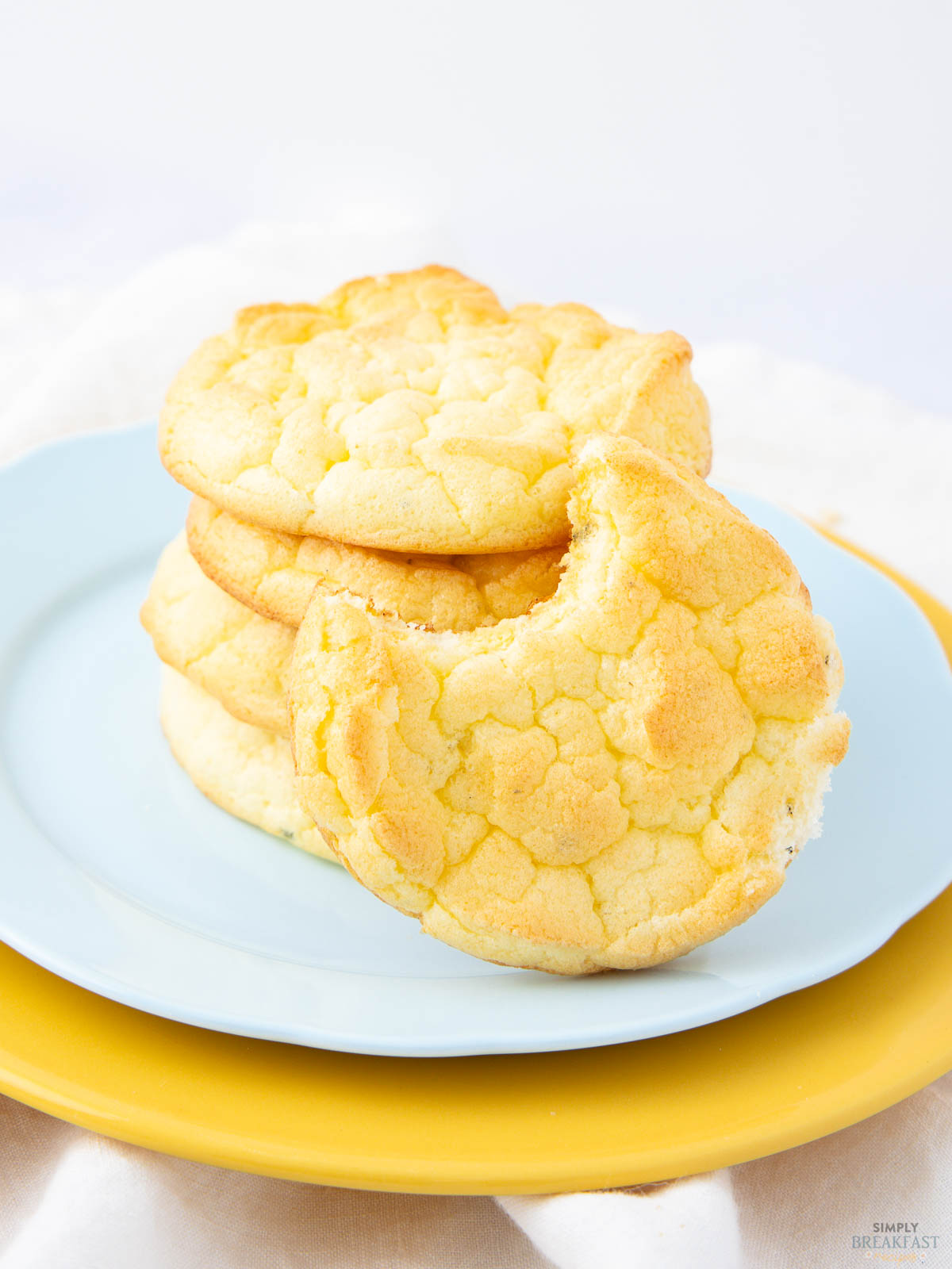 A stack of fluffy cookies with a golden crust on a light blue plate, placed on a yellow plate. The top cookie has a bite taken out of it, and the background is softly blurred, creating a bright, airy feel.