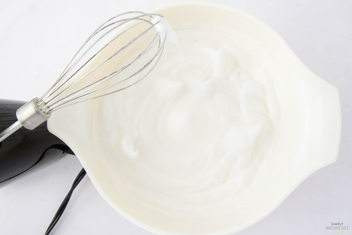 A mixing bowl with fluffy, whipped egg whites and a whisk attachment from an electric mixer is visible. The mixture appears light and airy, suggesting it has been beaten to peaks. The mixer is partially visible on the left.