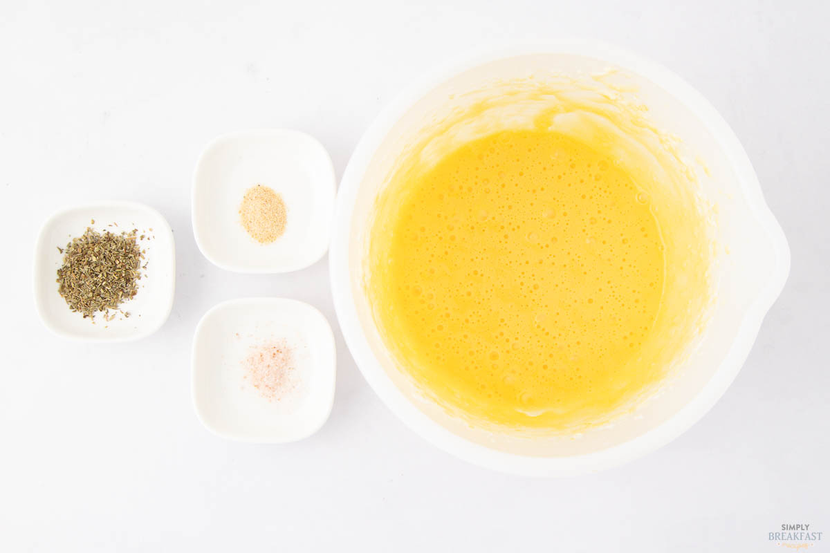 A white bowl filled with beaten eggs is placed next to three smaller square dishes containing black pepper, garlic powder, and pink salt on a white background.