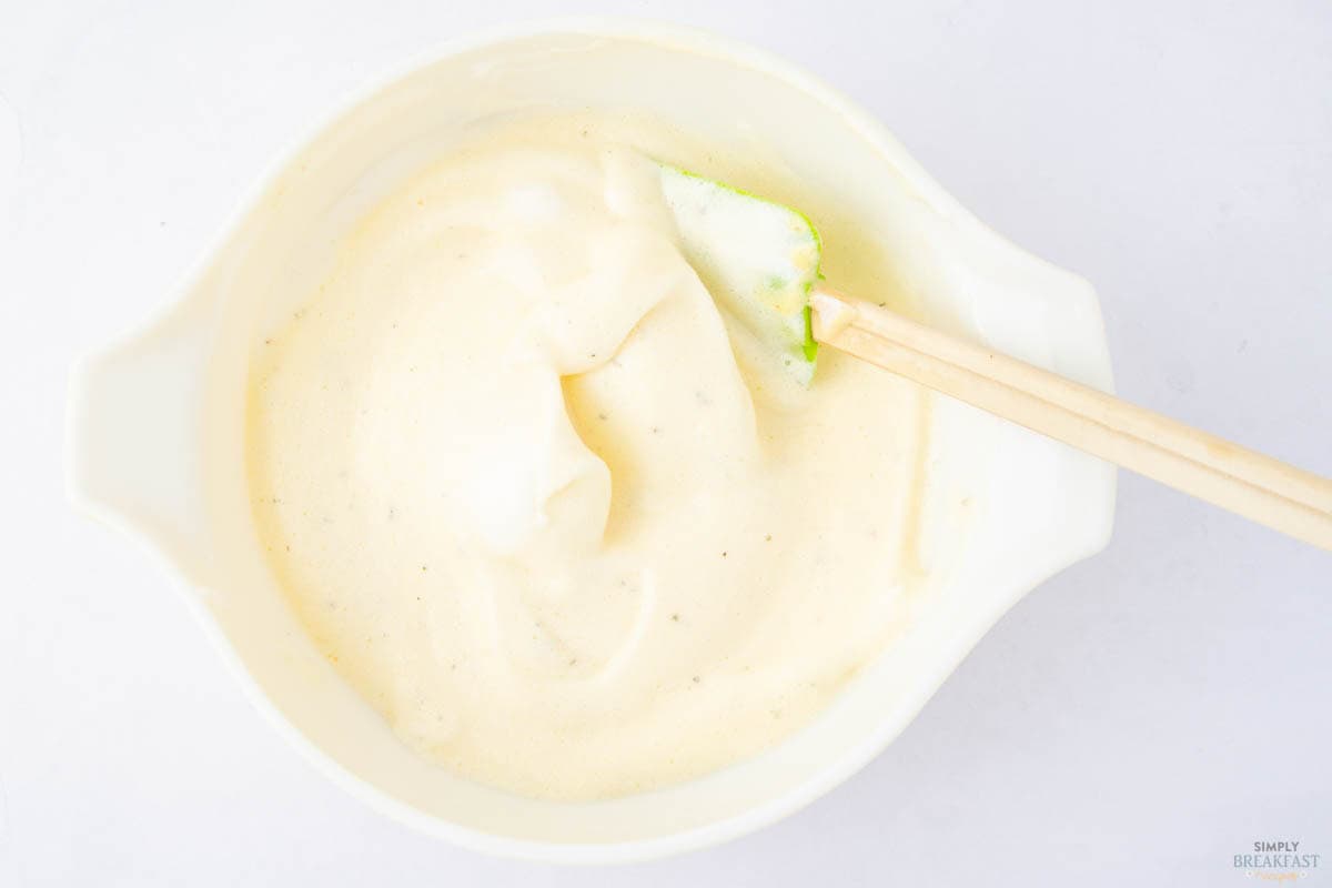 A white bowl containing whipped egg whites with soft peaks, and a light green spatula resting inside. The creamy mixture has a smooth texture. The background is a plain white surface.