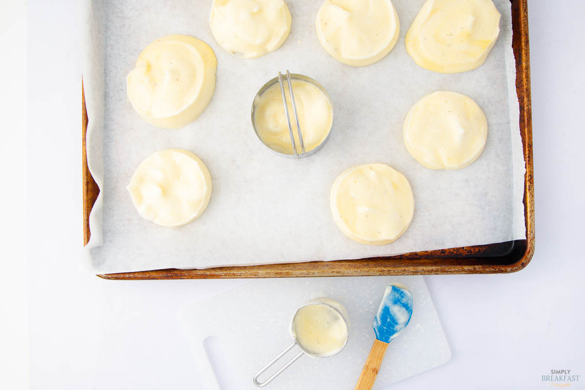 A baking tray holds eight uncooked biscuits or scones on parchment paper, arranged in a circular pattern. A round metal cutter is in the center. Below, a cutting board with spilled batter and a blue spatula are visible.