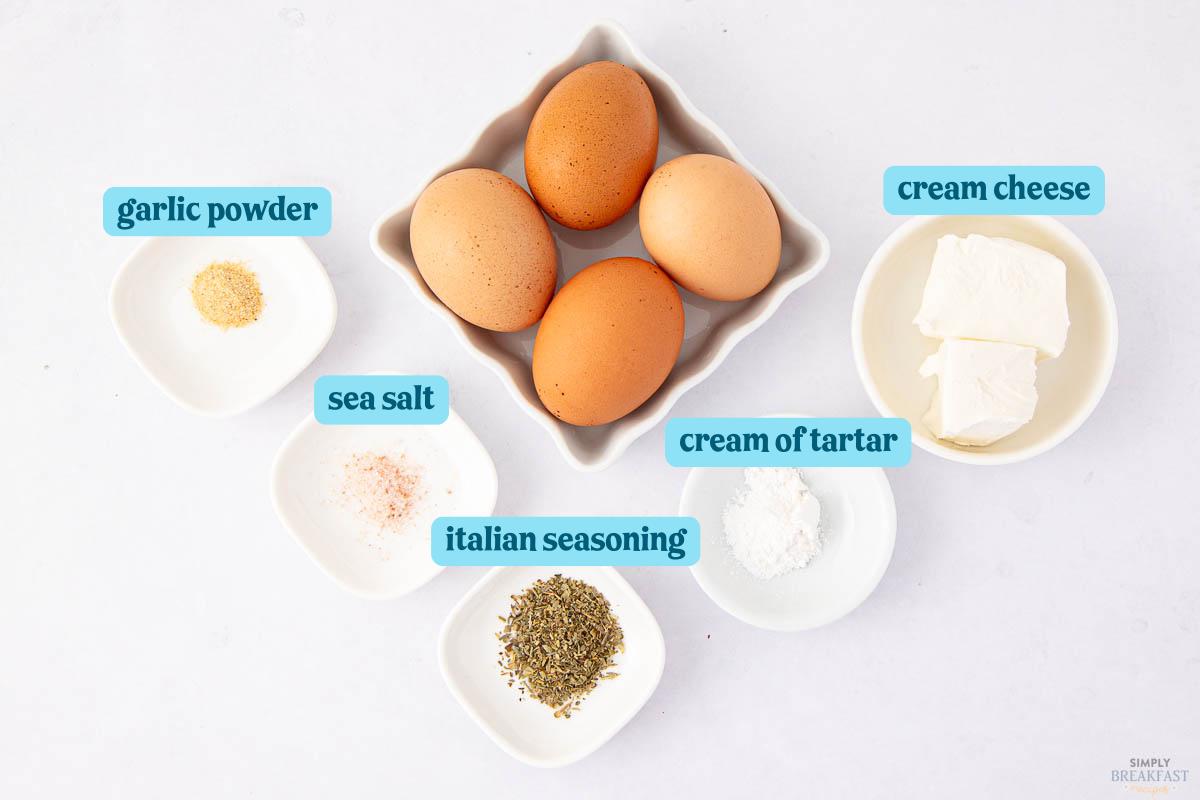 Image displays ingredients laid out for a recipe. There are eggs, cream cheese, garlic powder, sea salt, Italian seasoning, and cream of tartar in separate dishes on a white surface, each labeled with text.