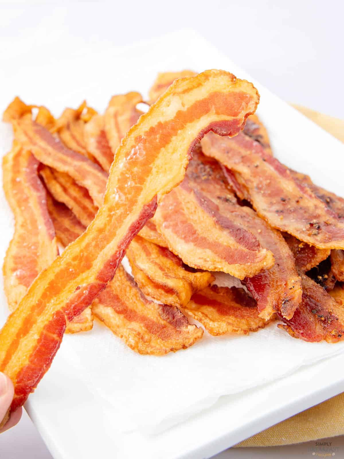 A hand holding a crispy strip of cooked bacon over a white plate stacked with more bacon strips. The bacon appears golden-brown and is arranged on a paper towel to absorb excess grease.