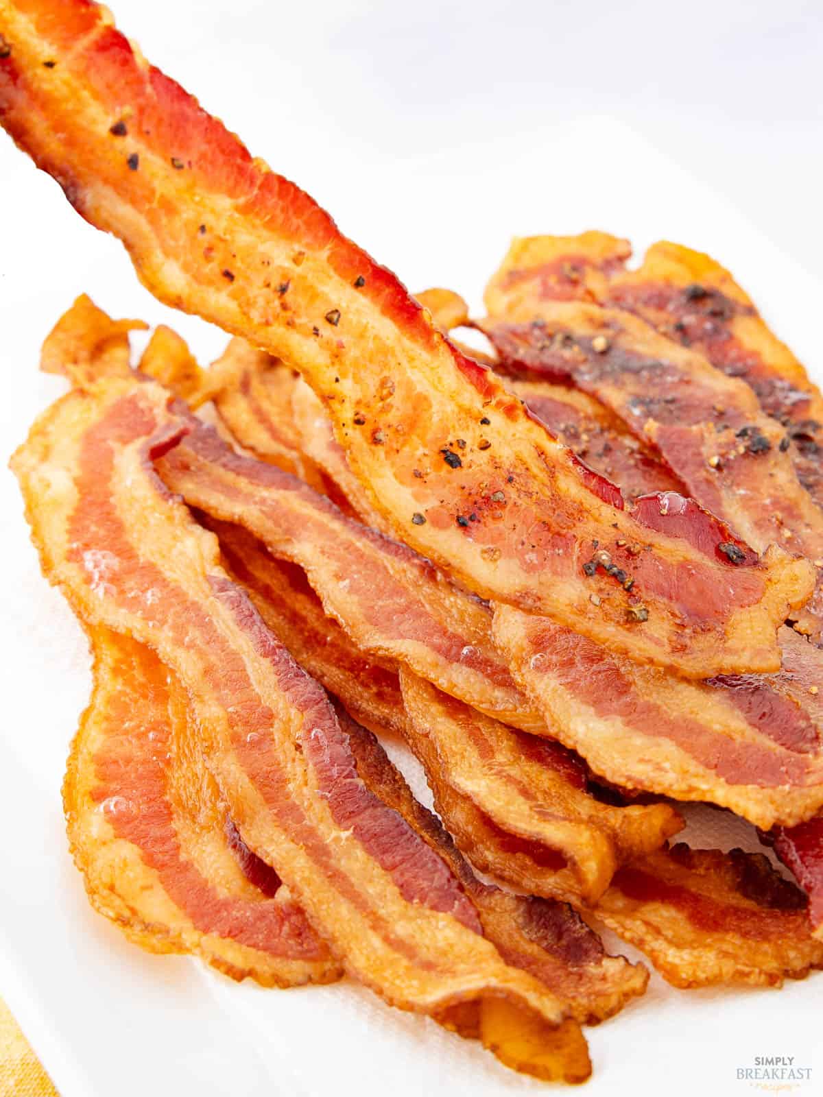 A close-up of several crispy, cooked bacon strips stacked on a white plate. One strip is lifted above the rest, showcasing its golden-brown color and textured surface.