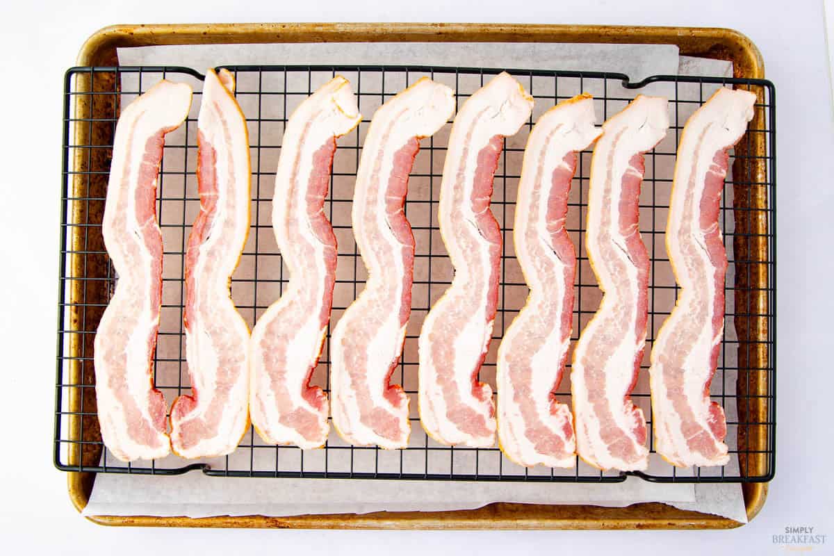 Strips of uncooked bacon are neatly arranged side by side on a metal rack placed over a baking sheet lined with parchment paper. The bacon is ready for baking.