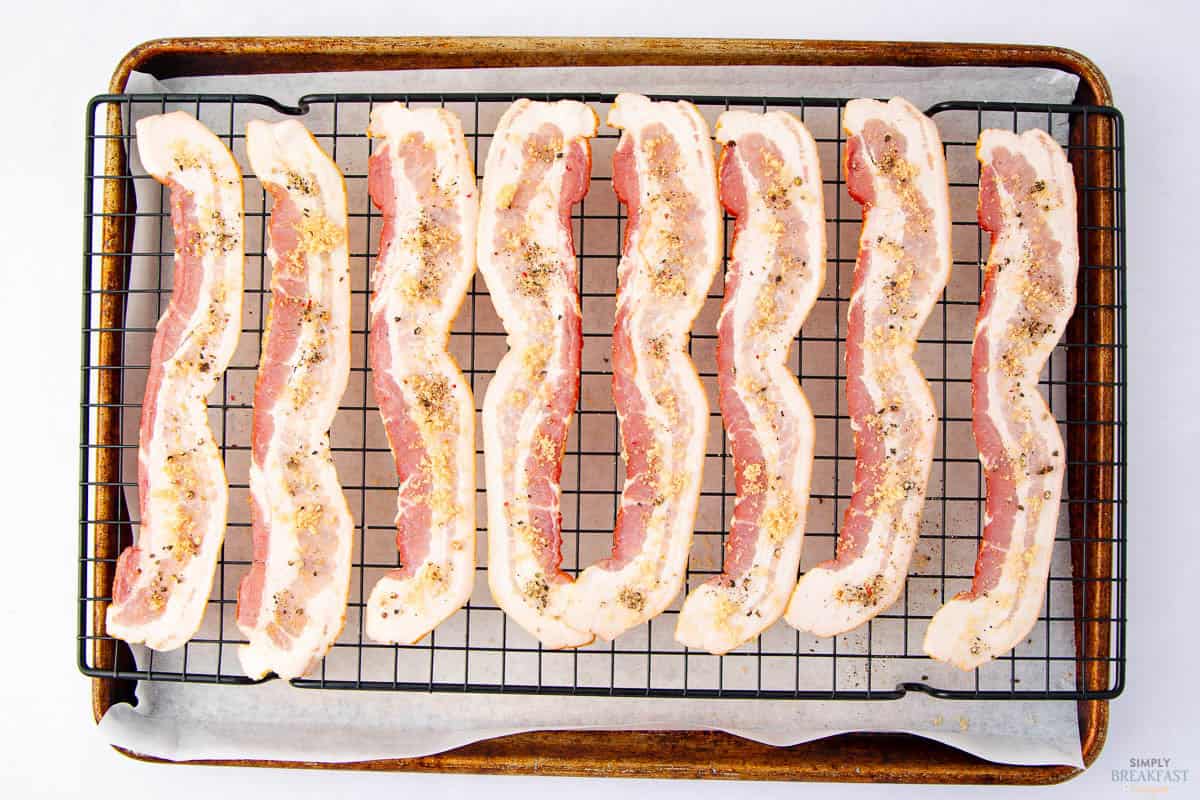 Strips of bacon seasoned with black pepper and spices are arranged in neat rows on a wire rack placed on a baking sheet lined with parchment paper.