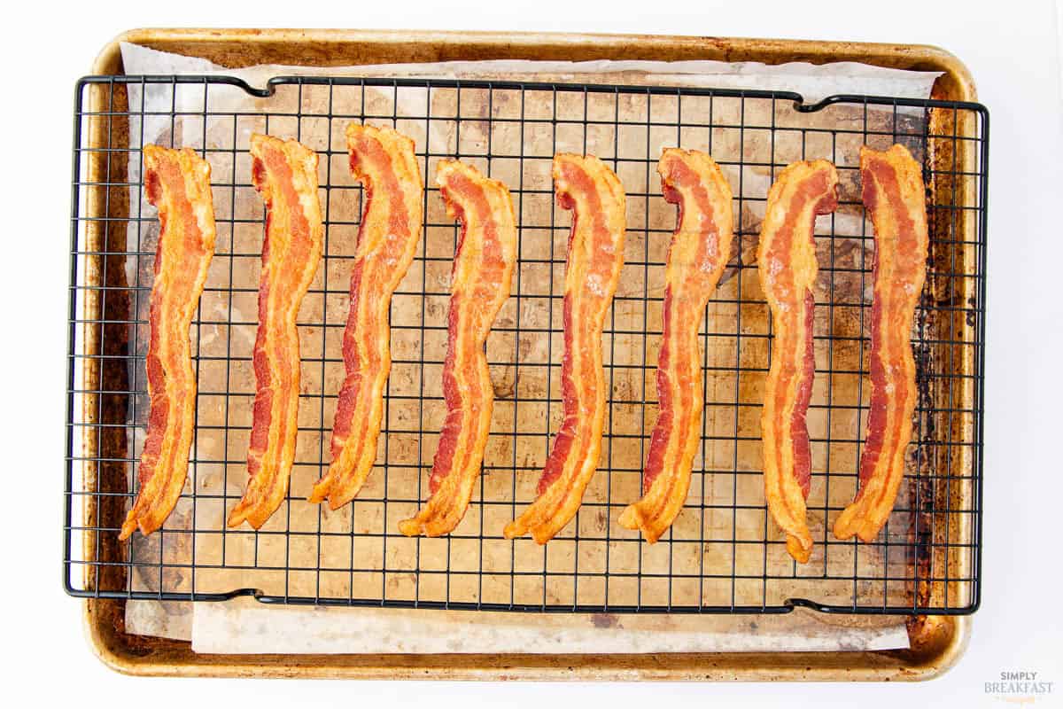 Eight crispy strips of bacon are lined up on a black wire rack placed on a baking sheet covered with parchment paper. The bacon is evenly spaced and golden brown.