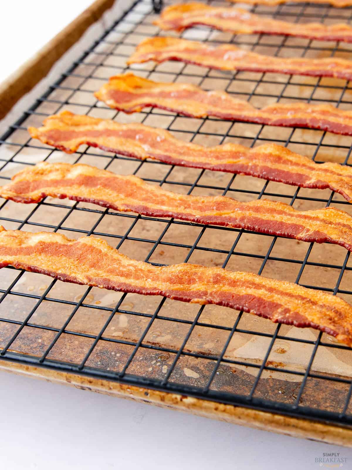 Crispy strips of bacon are evenly spaced on a cooling rack set over a baking sheet. The bacon appears well-cooked with a golden-brown texture.